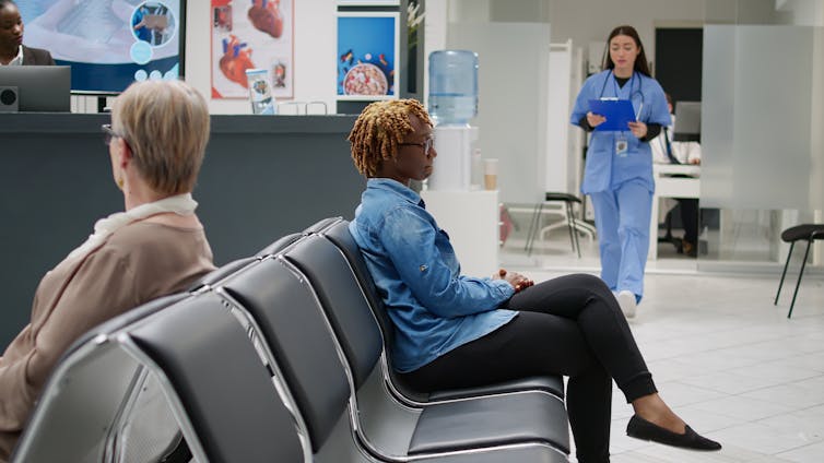 A clinic waiting room with two women sitting in chairs and a person in scrubs approaching