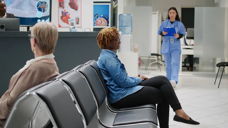 A clinic waiting room with two women sitting in chairs and a person in scrubs approaching