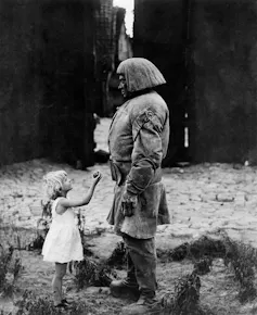 A black and white photo of a little girl in a white dress holding up a piece of fruit to a huge man in dirty clothes in an alleyway.