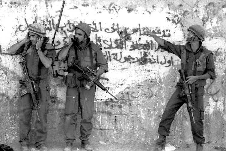 Three men in army clothing and armed stand by a wall.