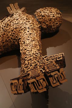 A human figure carved out of wood is positioned lying down, with intricate Hebrew letters carved into the surface.