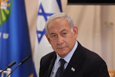 A middle aged man dressed in a business suit stands near a white flag with a blue star.