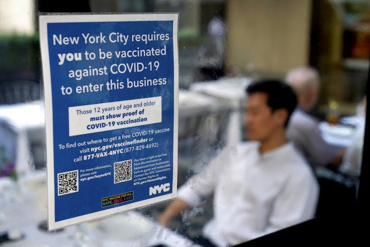 Sign on window reading 'New York City requires you to be vaccinated against COVID-19 to enter this business,' with a person sitting at a desk inside the room