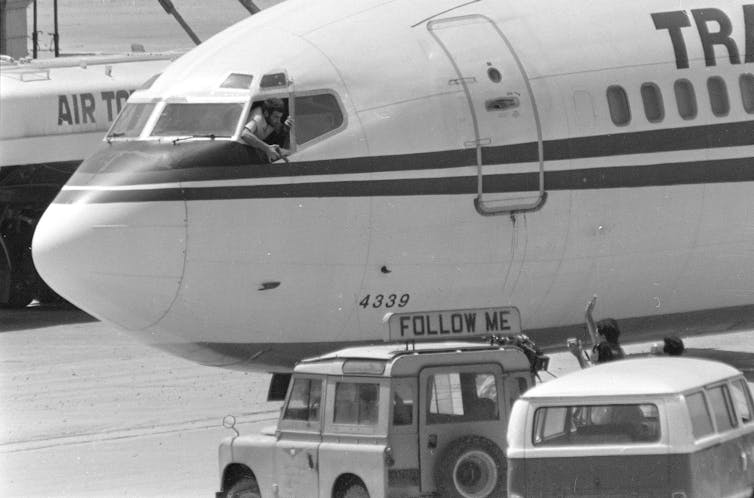 A man pokes his head out of the cockpit of a jet and points a pistol at a news crew.