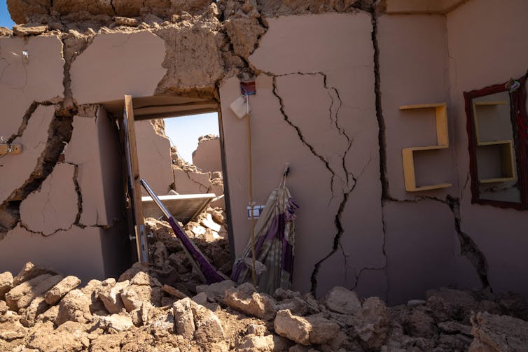 The few standing walls of a house are severely cracked. The roof is gone, and rubble lies on the floor.