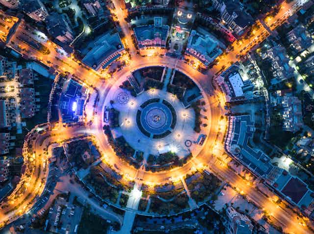 A birds-eye view of a traffic circle at night in a cirty center, around a circular pavilion, with 7 roads going off of the circle. The streets are lit in yellow light.