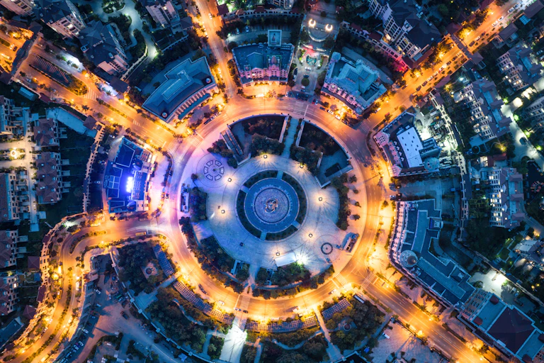 A birds-eye view of a traffic circle at night in a cirty center, around a circular pavilion, with 7 roads going off of the circle. The streets are lit in yellow light.