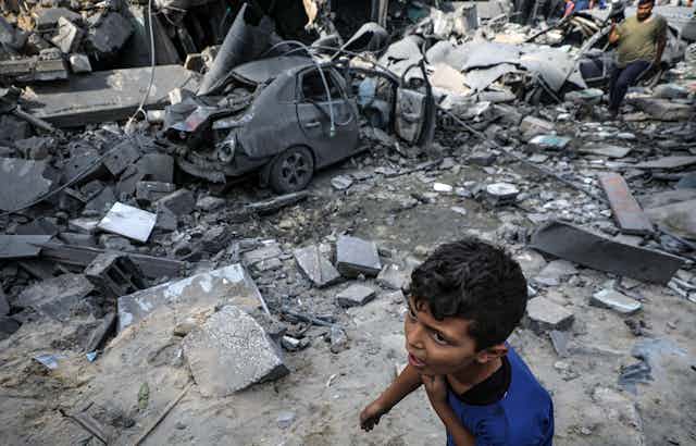 Um menino de camisa azukl ao lado de um automóvel destruído por bombardeio