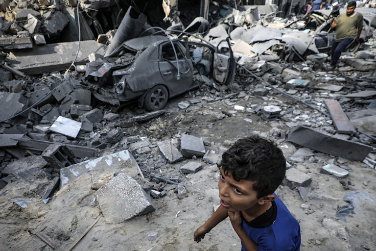 Um menino de camisa azukl ao lado de um automóvel destruído por bombardeio