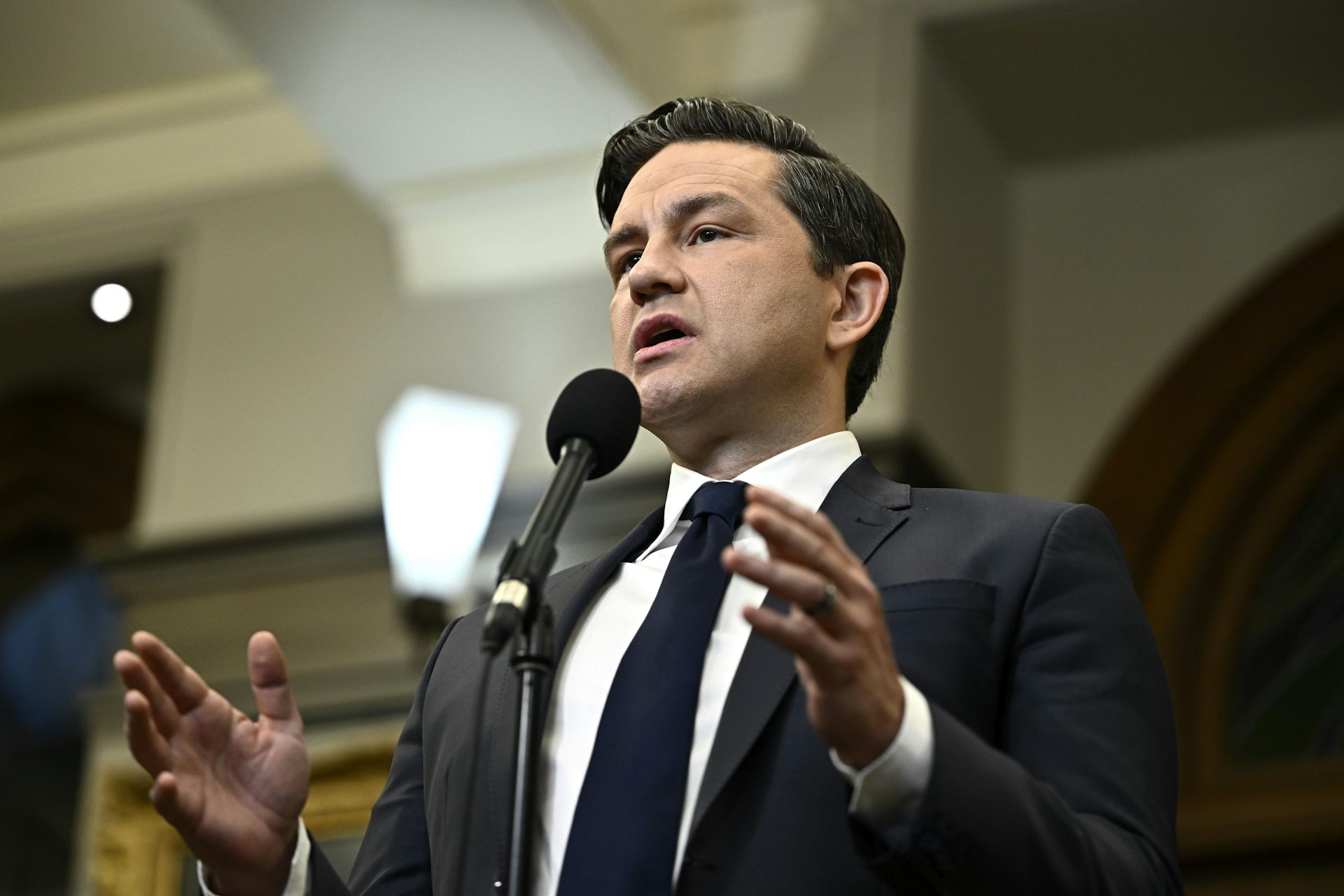 A dark-haired man gestures while speaking into a microphone.