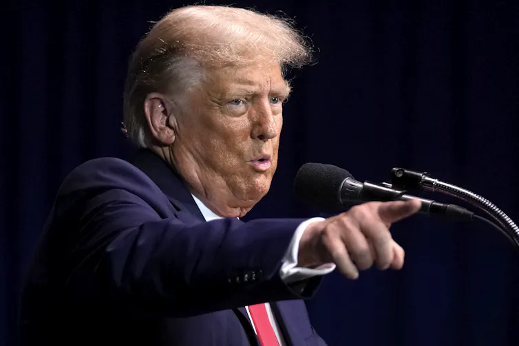 A blond older man gestures while speaking into a microphone.