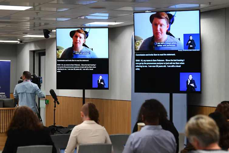 television screens show a woman with disability speaking, seated people listen