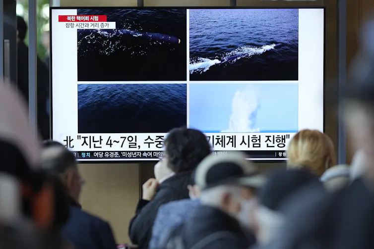A photo of people watching a large TV showing images of explosions at sea accompanied by text in Korean.
