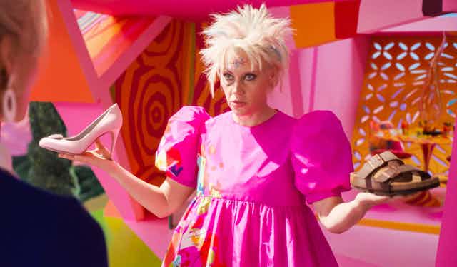 Kate McKinnon as Weird Barbie in the movie holding a pink stiletto and a brown Birkenstock sandal.