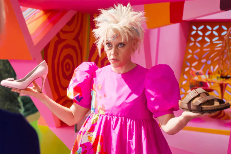 Kate McKinnon as Weird Barbie in the movie holding a pink stiletto and a brown Birkenstock sandal.