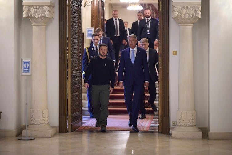 Volodymyr Zelenskyy walks down steps and through a large door with other men in suits and uniform.