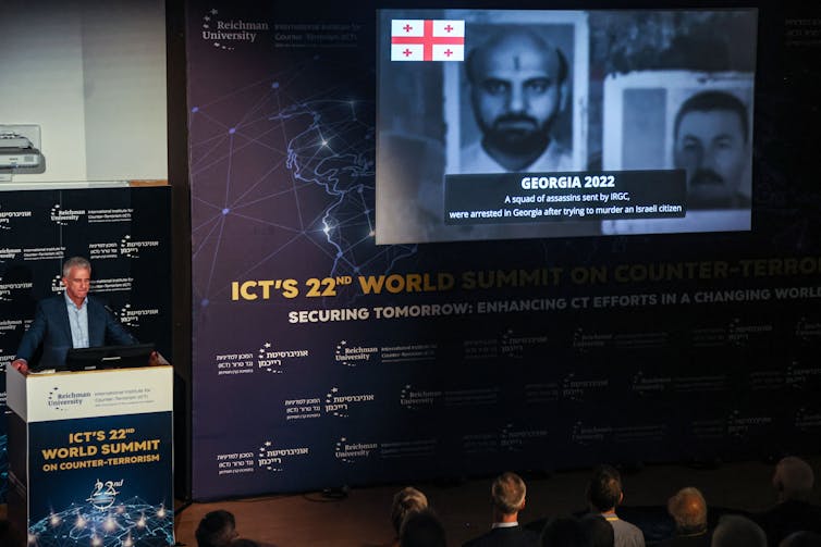 A man with a suit stands at a podium that says 'ICT's 22nd world summit on counter-terrorism' and next to a large screen that shows headshots of people