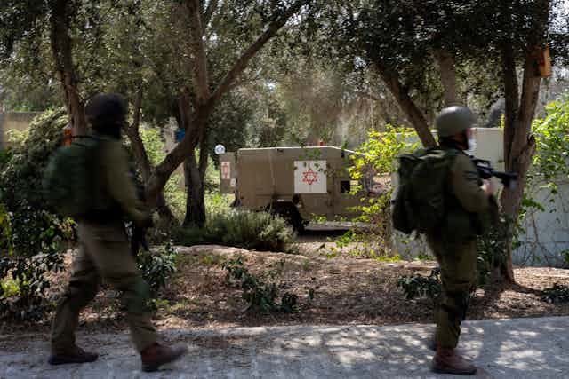 Two soldiers in green hold guns and walk past a medical vehicle with a Jewish star on it.