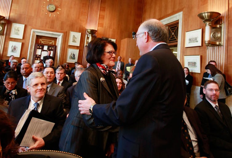 A woman and man shake hands in a crowded hearing room.
