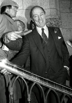 Black and white photo of Pierre Trudeau smiling and holding a railing, surrounded by reporters