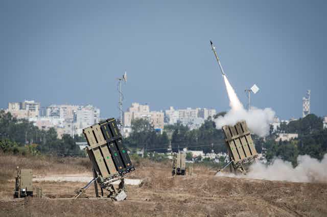 a missile with smoke trailing behind it emerges from one of two box-like structures in a field with a city skyline in the background