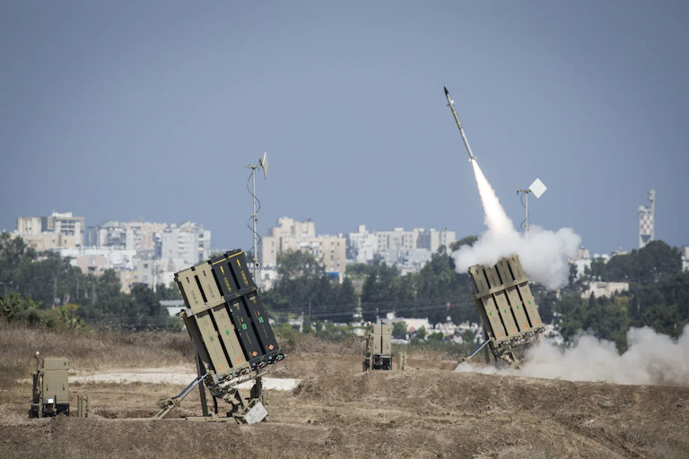 a missile with smoke trailing behind it emerges from one of two box-like structures in a field with a city skyline in the background