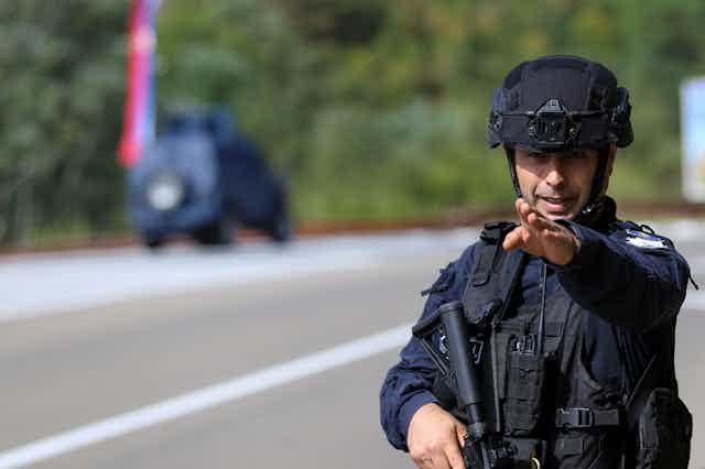 A heavily armed police officer holds up his hand while guarding a road.