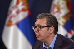 A man with dark hair and glasses in profile with white blue and red flags behind him.