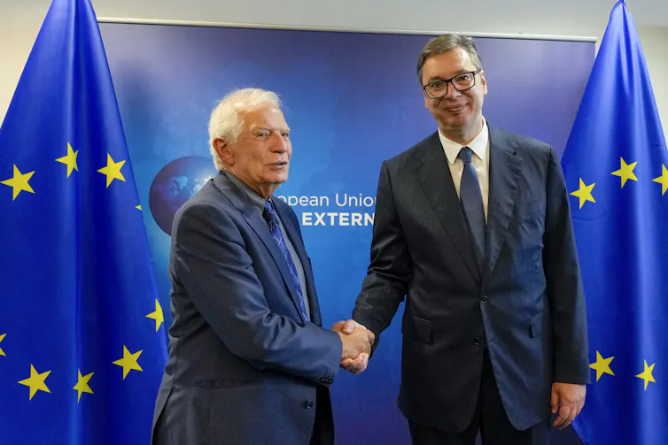 Two men, a tall dark-haired man and a shorter white-haired man, shake hands.