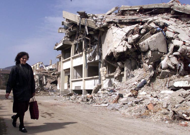 A woman in dark clothing walks past a destroyed building.