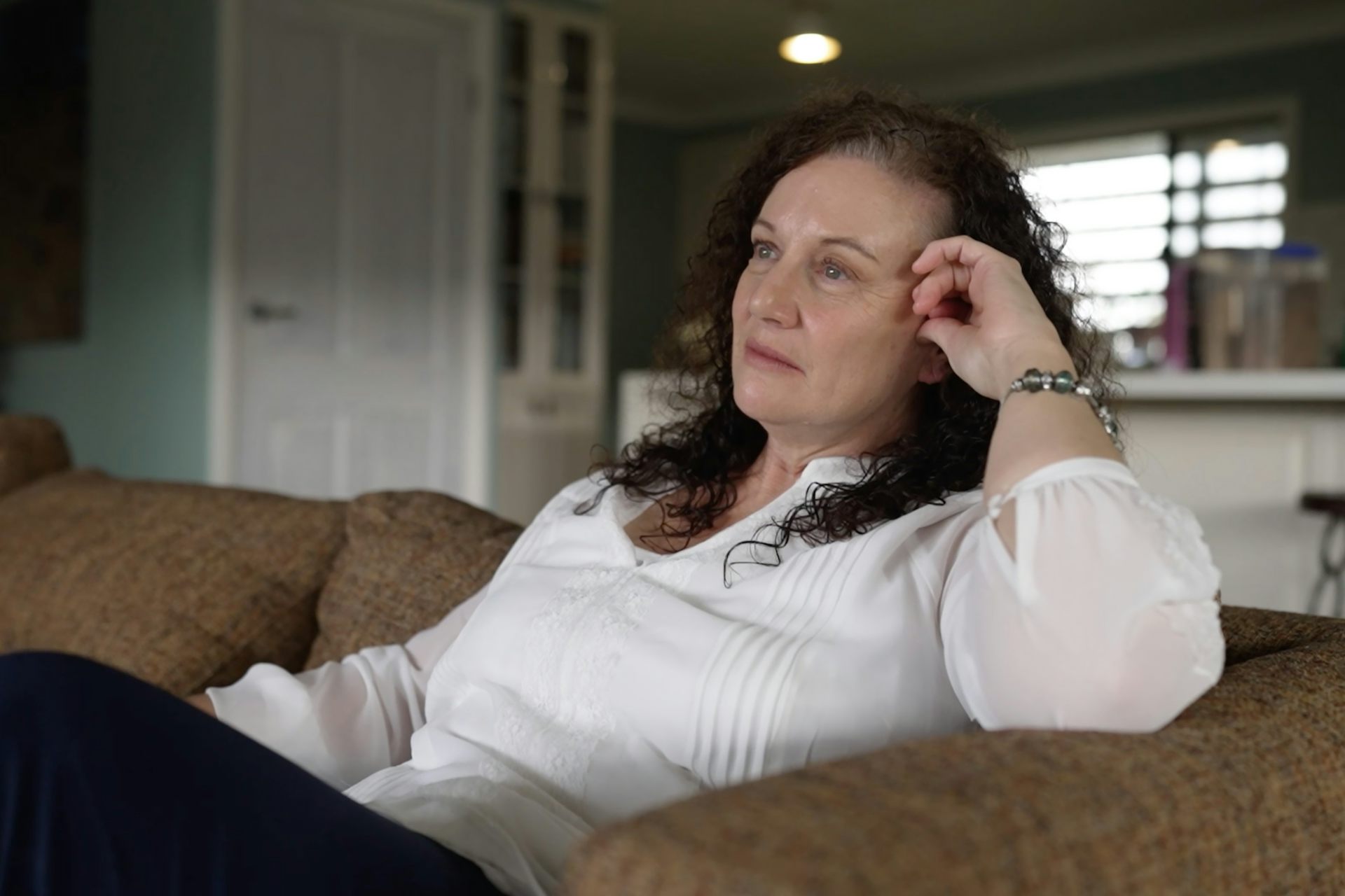 A woman in a white shirt sits on a couch and looks into the distance
