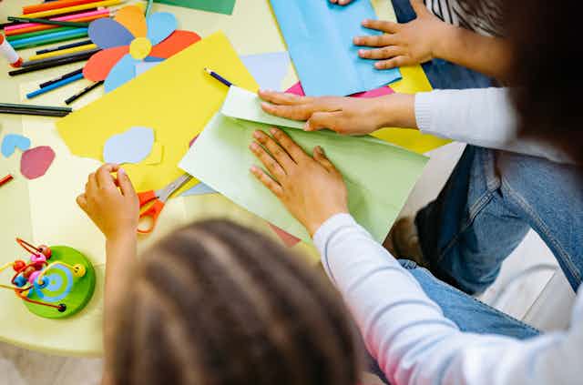 A teacher and children work with coloured paper, pencils and scissors.