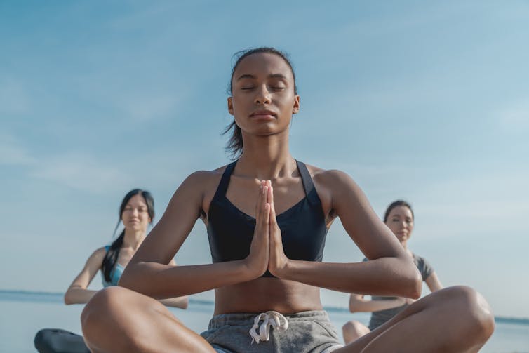 Trois jeunes femmes, l’une devant, les deux autres derrière sont assises en tailleur, les mains jointes sur le cœur dans une position de yoga.