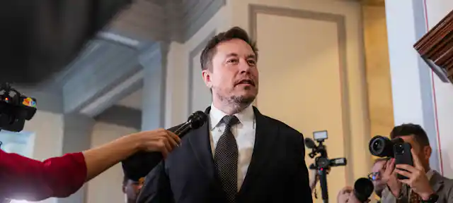 Elon Musk speaking to reporters, people with cameras and microphones.