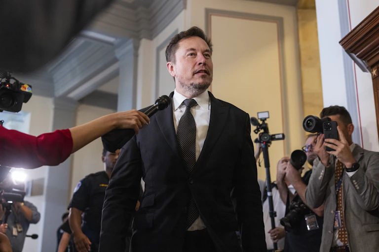 Elon Musk speaking to reporters, people with cameras and microphones.