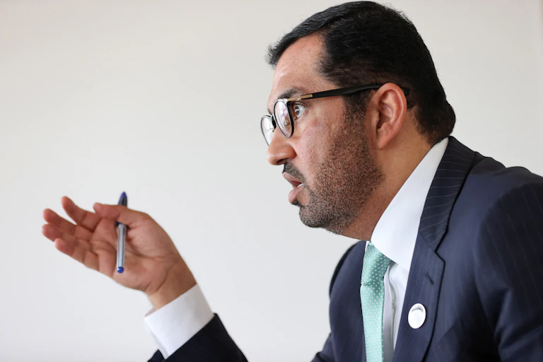 A side profile photo shows a man in a western business suit gesturing with his hand while holding a pen and looking intently at something off camera to the left as he speaks.