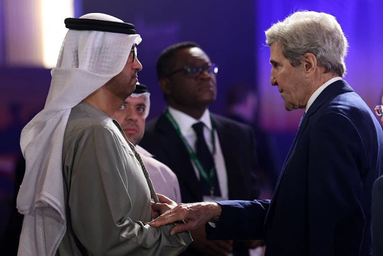 Kerry, in a western business suit, touches Al Jaber's arm as they speak. Al Jaber is in traditional Middle Eastern attire. Both men are tall and about the same height.