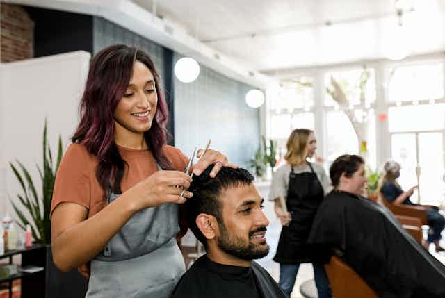 Hairdresser talks to client while cutting hair, other hairdressers and customers in the background.