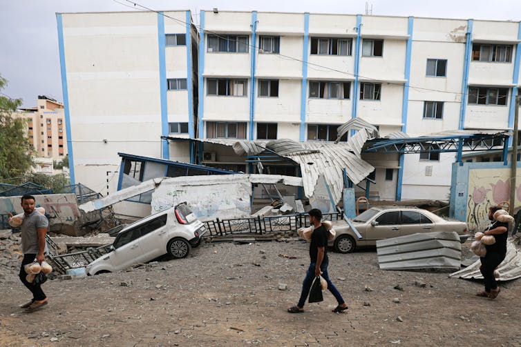 Three men walk past a dilapidated looking building and cars that have been partially destroyed and fallen into a crater in the ground.
