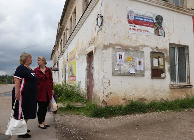 Two women carrying bags stand next to a decrepit building.