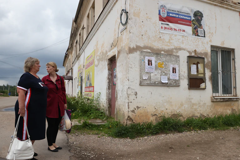 Two women carrying bags stand next to a decrepit building.