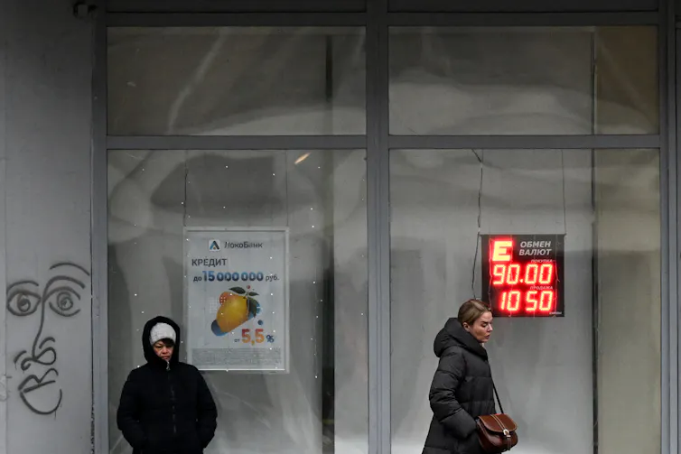 Two people stand outside a glass building with currency figures in neon.