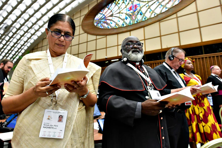 A woman in a white outfit and glasses stands next to a man in a black clerical outfit with a white beard as they read from a program during a service.