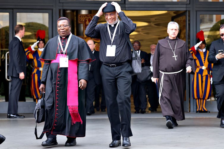 A man in a black clerical robe with a pink sash and another man in all black, both wearing name tags, smile as they leave a building.