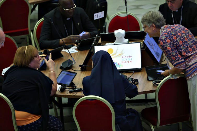 Three women, one of whom wears a headcovering, chat at a round table as three men look at laptops and phones.