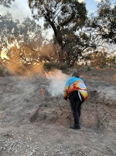 A person in colourful clothing wafting smoke over a mud bank with rising sun in the background