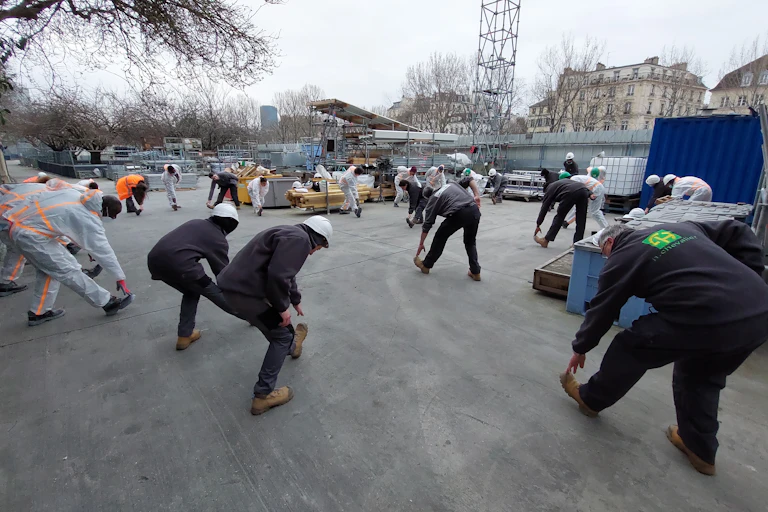 Sur un chantier du BTP, des travailleurs sont penchés en avant. Ils sont en train de pratiquer des exercices physiques.
