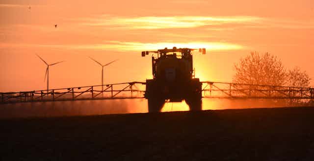 Un tracteur épand du glyphosate sur un champ, à l'aube, devant un paysage d'éoliennes