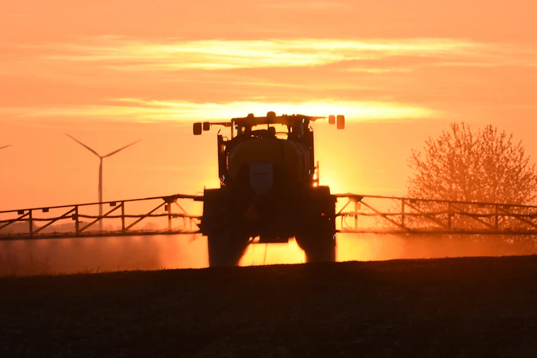 Un tracteur épand du glyphosate sur un champ, à l'aube, devant un paysage d'éoliennes