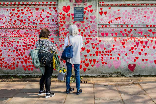 Two people stand in front of a wall covered in red hearts.